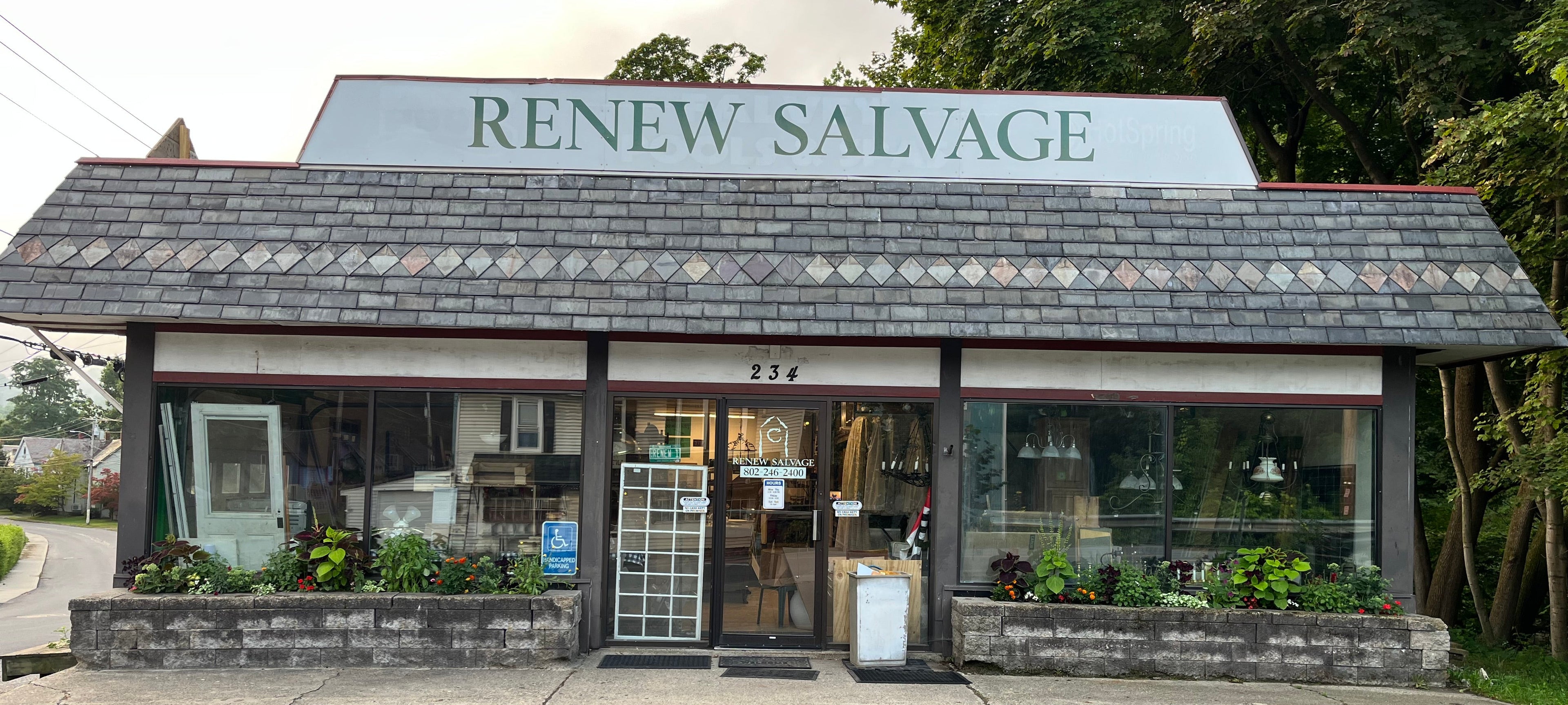 Front exterior of a small salvage shop named RENEW SALVAGE, with a large sign above the entrance, glass windows, a gray shingled roof, and trees behind the building.
